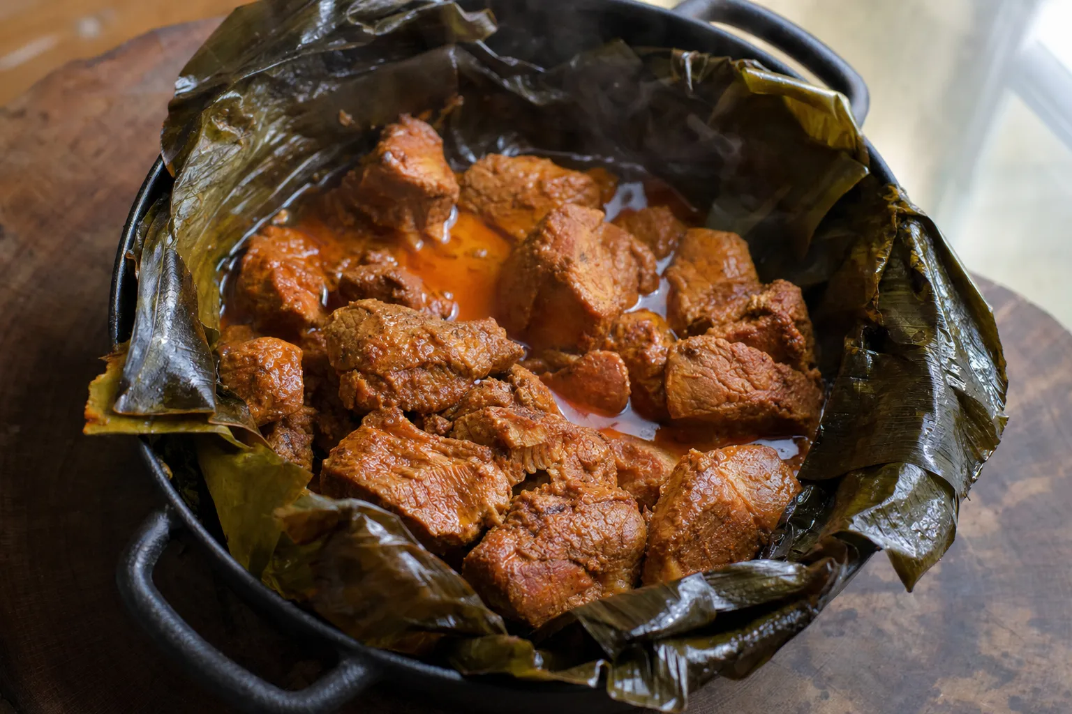 ダッチオーブンの中でバナナの葉を開いた、オレンジ色の汁に浸かった焼き上がりの角切りの豚肉
