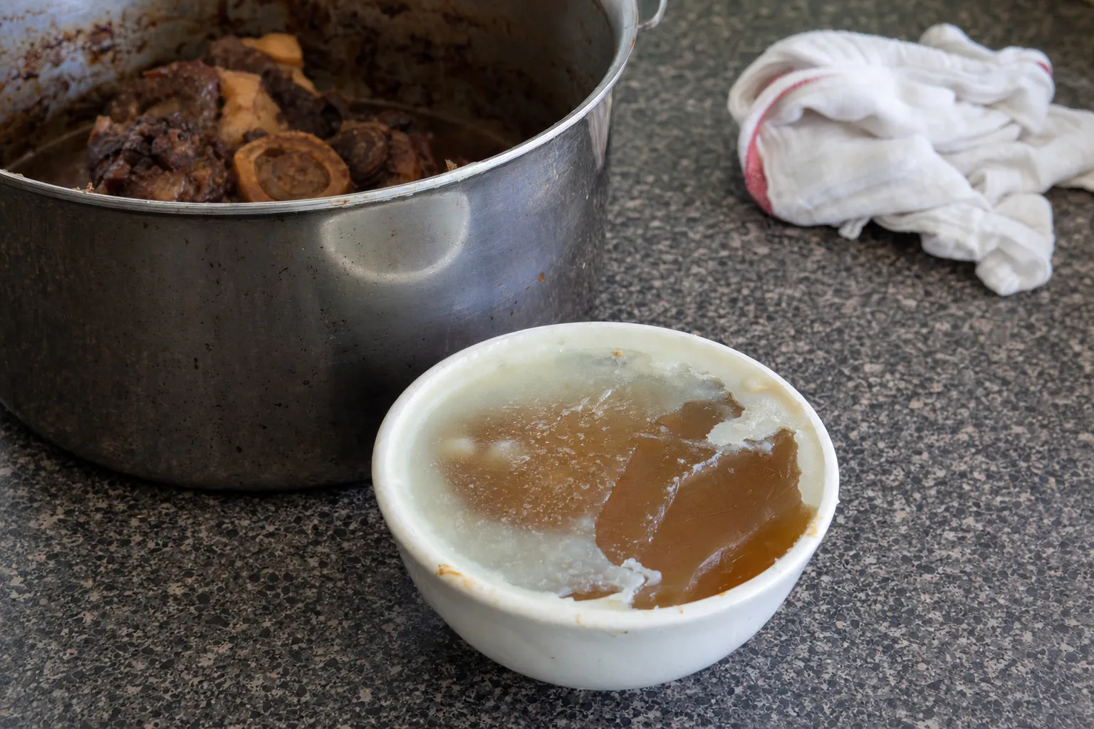 白い脂の層の下で琥珀色のゼリー状に固まった煮汁の器と、奥に骨付きのすね肉が入った鍋